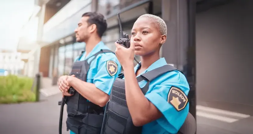 Two uniformed officers, a woman using a walkie-talkie and a man standing beside her, maintain a professional stance outside a modern building, representing the career paths available to criminal justice graduates.